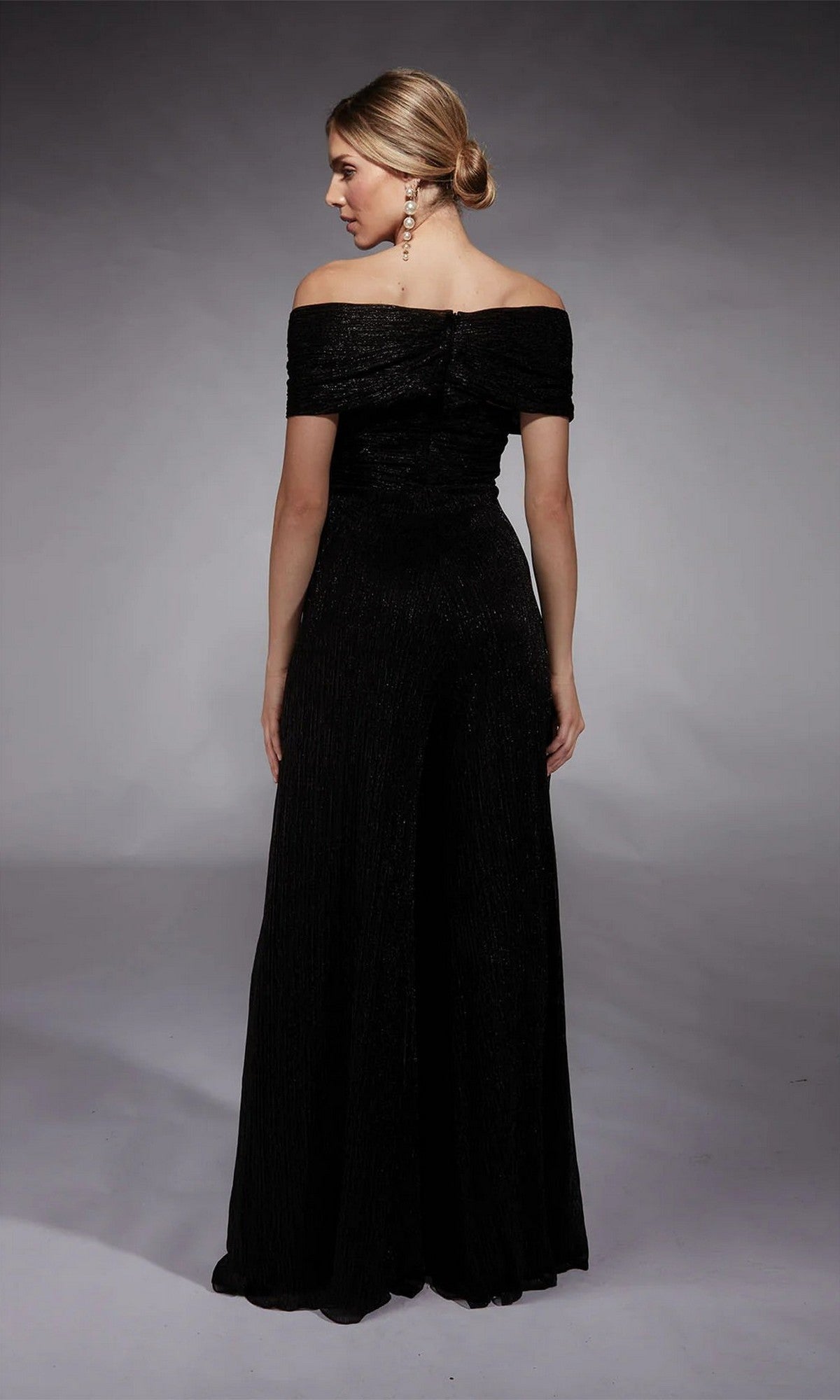 A woman faces away, her hair in a low bun, wearing the Alyce 27768 long formal off-the-shoulder black crinkle chiffon jumpsuit.